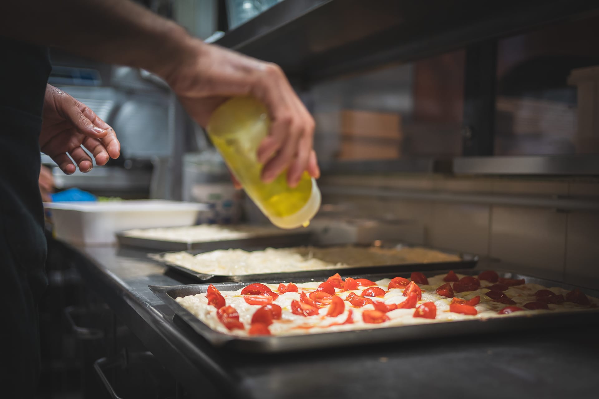 mano del cuoco che versa olio extravergine di oliva su teglia di focaccia con pomodorini